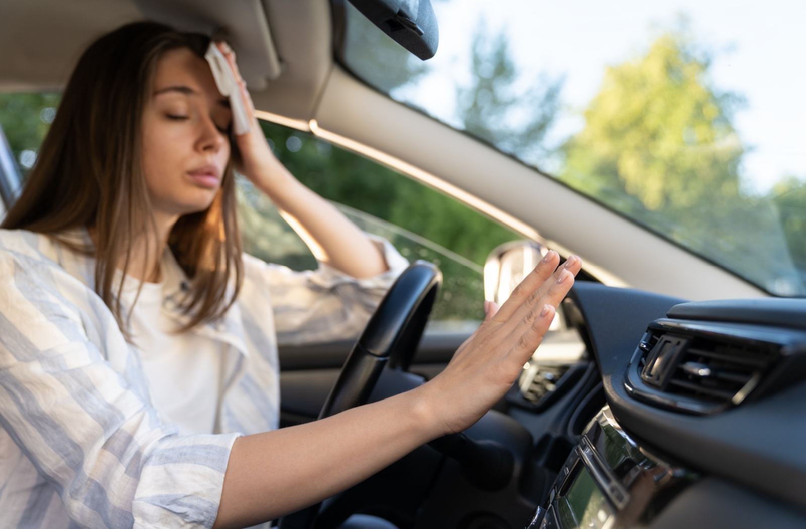 A woman dabbing her face with a napkin while feeling hot air from her car's AC system.
