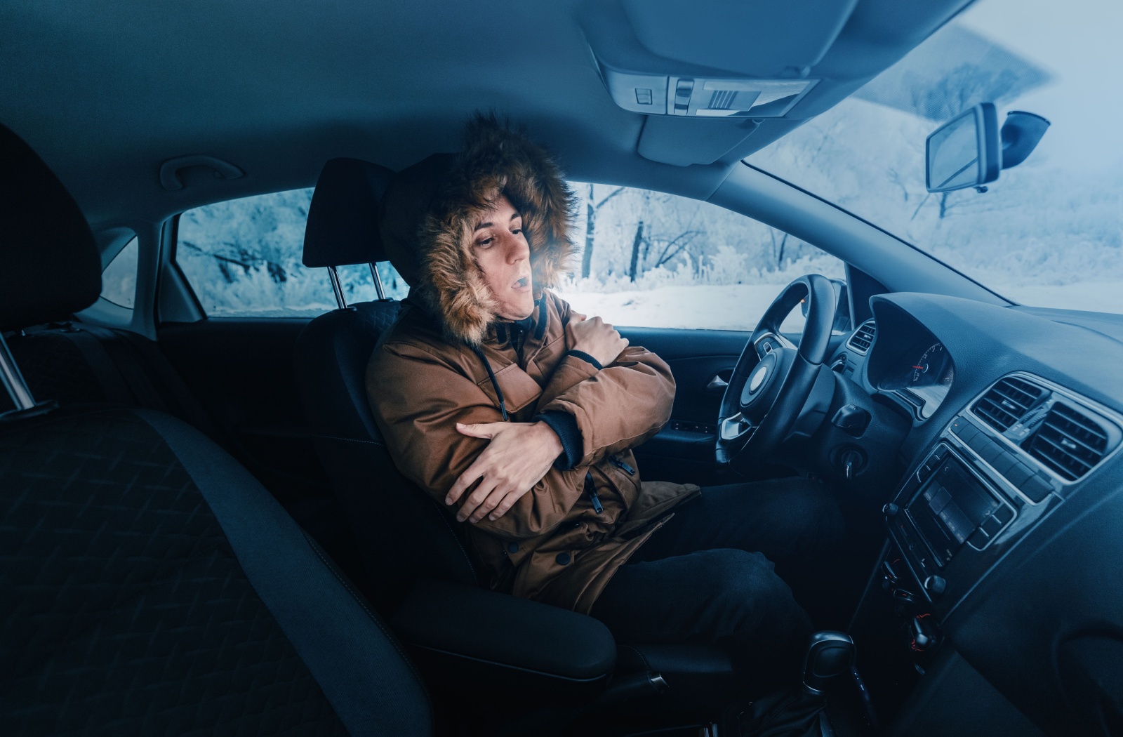 Person wearing a winter coat, sitting inside a cold car and shivering, with visible breath in the frosty air.