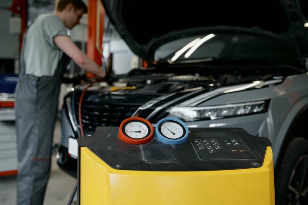 A mechanic working under the hood of a vehicle in a repair shop, with an A/C service machine and pressure gauges in the foreground.