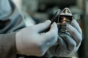 A person in gloves examines a metal thermostat component closely
