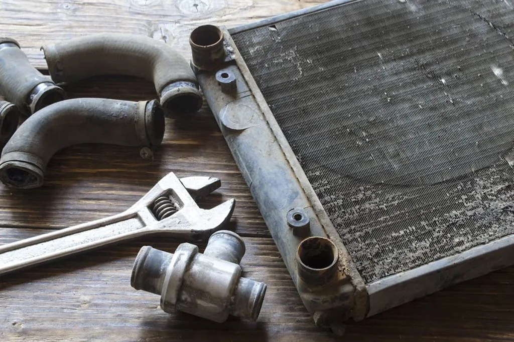 A disassembled radiator sits alongside worn hoses and a wrench on a wooden surface